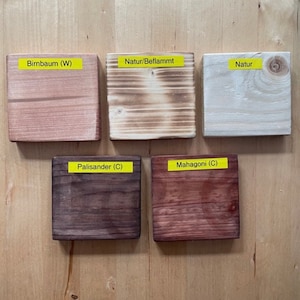 May include: Five wooden blocks with different finishes and colors are displayed on a light wood surface. The blocks have labels that read "Birnbaum (W)", "Natur/Beflammt", "Natur", "Palisander (C)", and "Mahagoni (C)".