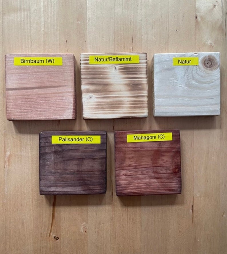 May include: Five wooden blocks with different finishes and colors are displayed on a light wood surface. The blocks have labels that read "Birnbaum (W)", "Natur/Beflammt", "Natur", "Palisander (C)", and "Mahagoni (C)".