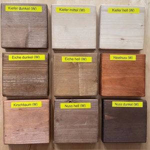 May include: Nine wooden blocks with different wood stain colors, including dark, medium, and light tones. Each block has a yellow label with German text. The colors range from light beige to dark brown.