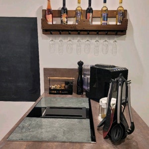 May include: A rustic wooden wine rack with six wine bottles and eight wine glasses hanging below. The rack is mounted on a white wall above a countertop with a built-in cooktop and various kitchen utensils.