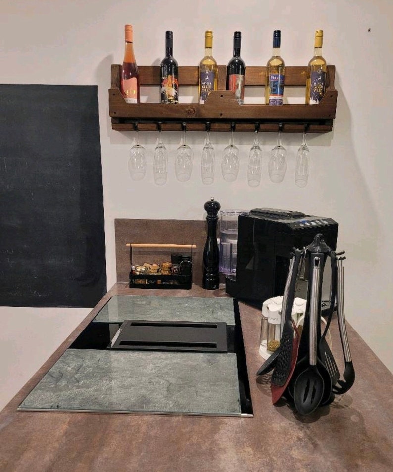 May include: A rustic wooden wine rack with six wine bottles and eight wine glasses hanging below. The rack is mounted on a white wall above a countertop with a built-in cooktop and various kitchen utensils.