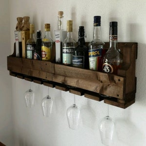 May include: A rustic, dark-stained wooden wine rack mounted on a white wall. The rack holds various liquor bottles and has slots for hanging wine glasses. Three clear wine glasses are suspended below the rack, showcasing the design.