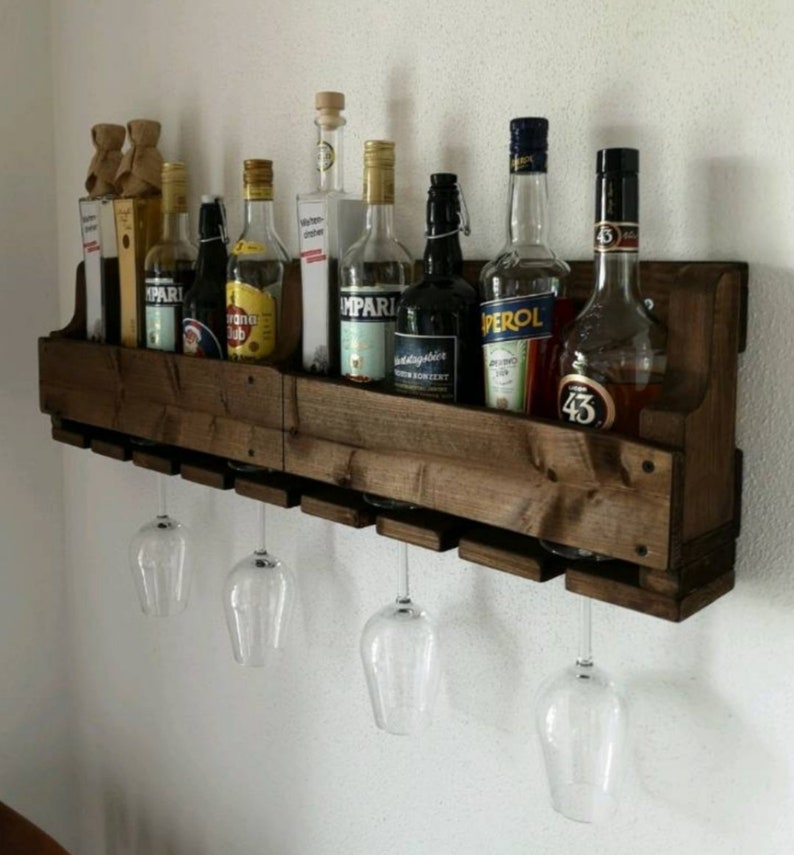 May include: A rustic, dark-stained wooden wine rack mounted on a white wall. The rack holds various liquor bottles and has slots for hanging wine glasses. Three clear wine glasses are suspended below the rack, showcasing the design.