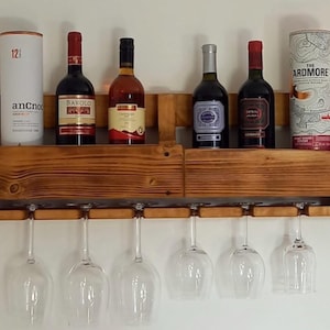 Può includere: Un portabottiglie in legno con un ripiano per bottiglie e fessure per appendere i calici. Il portabottiglie contiene diverse bottiglie di vino e un contenitore cilindrico. Sei calici sono appesi sotto. Il legno ha una finitura calda e naturale.