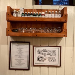 Peut inclure: Une étagère en bois à deux niveaux, remplie de petites bouteilles en verre et de verres. L'étagère est fixée à un mur blanc. Il y a deux images encadrées sur le mur sous l'étagère. L'image du haut porte le texte "The Classic Core Range Flight" et l'image du bas présente un schéma du processus de distillation.