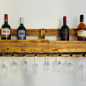 May include: A rustic, wooden wine rack with a natural finish, holding four wine bottles and eight wine glasses. The rack is mounted on a white wall, showcasing the bottles and glasses.