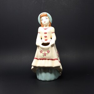 Puede incluir: Una figurita de cerámica de una mujer con un vestido azul y blanco con flores rojas. Ella sostiene una cesta en sus manos. La figurita está pintada en un estilo vintage.