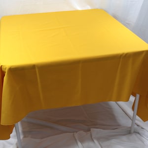 Puede incluir: Un mantel cuadrado de color amarillo brillante drapeado sobre una mesa. El mantel está hecho de una tela lisa y sin arrugas.