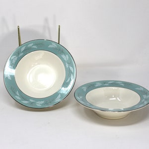 May include: Two white ceramic bowls with a teal and silver leaf design. The bowls are empty and sitting on a white surface.