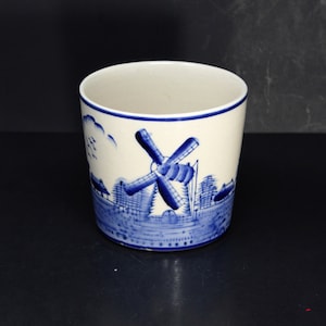 May include: A white ceramic mug with a blue and white windmill design. The mug features a windmill with a blue roof and white blades, surrounded by blue trees and a blue sky.
