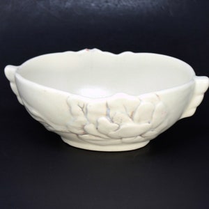 Puede incluir: Un tazón de cerámica blanco con un diseño floral. El tazón tiene una forma redondeada y un borde ancho. El diseño floral está en relieve en el exterior del tazón.