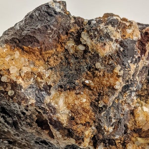 May include: A close-up of a rough, dark brown rock with embedded clear quartz crystals. The rock has a layered appearance with patches of orange and white.