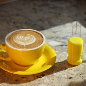 May include: A yellow ceramic coffee cup with a heart-shaped latte art design, sitting on a matching saucer. A yellow plastic holder with four thin metal needles is on the table next to the cup.