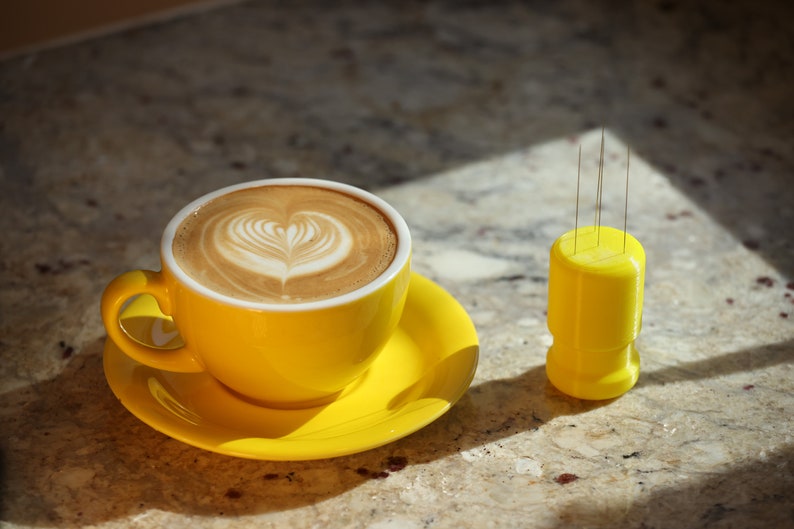 May include: A yellow ceramic coffee cup with a heart-shaped latte art design, sitting on a matching saucer. A yellow plastic holder with four thin metal needles is on the table next to the cup.