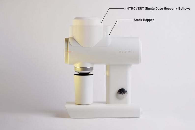 May include: A white coffee grinder with the text "sculptor..." and labels for "INTROVERT Single Dose Hopper + Bellows" and "Stock Hopper." The grinder has a cylindrical base and a cup-like container below.