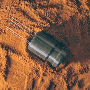 May include: A black and silver electronic component with multiple thin wires extending from the top. The component is sitting on a bed of brown powder.