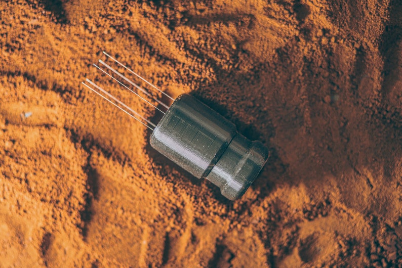 May include: A black and silver electronic component with multiple thin wires extending from the top. The component is sitting on a bed of brown powder.