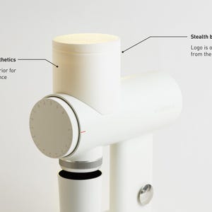 May include: A white kitchen appliance with a cylindrical design. It features a textured exterior for visual coherence and stealth branding, with the logo only visible from the back. The appliance has a dial with markings and a small, circular button.