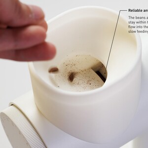 May include: Close-up of a white coffee grinder with a reliable anti-popcorn design. The text says the beans and fragments stay within the hopper and flow into the grinder for both slow feeding and full dosing. A hand is reaching towards the grinder.
