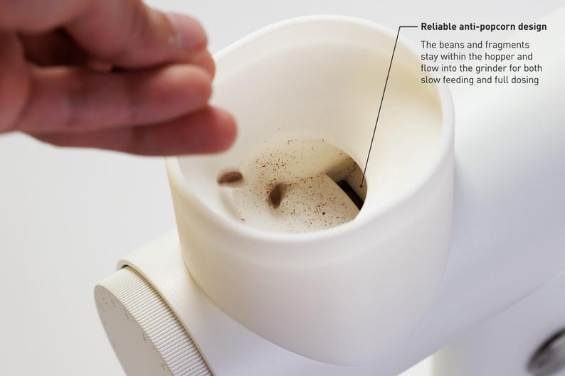 May include: Close-up of a white coffee grinder with a reliable anti-popcorn design. The text says the beans and fragments stay within the hopper and flow into the grinder for both slow feeding and full dosing. A hand is reaching towards the grinder.
