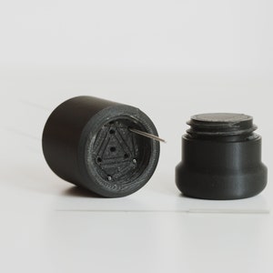 May include: A black plastic container with a threaded lid. The container has a triangular pattern with holes and a small metal pin sticking out.