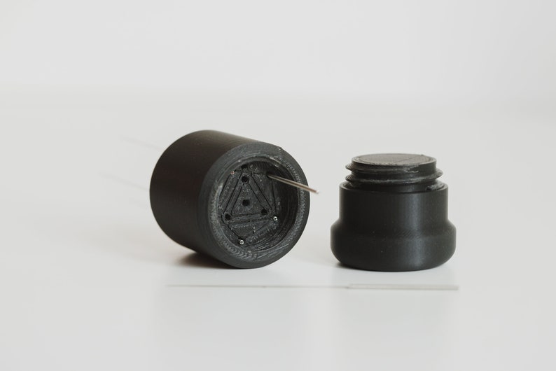 May include: A black plastic container with a threaded lid. The container has a triangular pattern with holes and a small metal pin sticking out.
