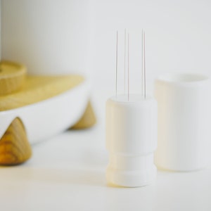 May include: A white ceramic holder with multiple thin needles sticking out of the top. The holder is sitting on a white surface next to another white ceramic holder.