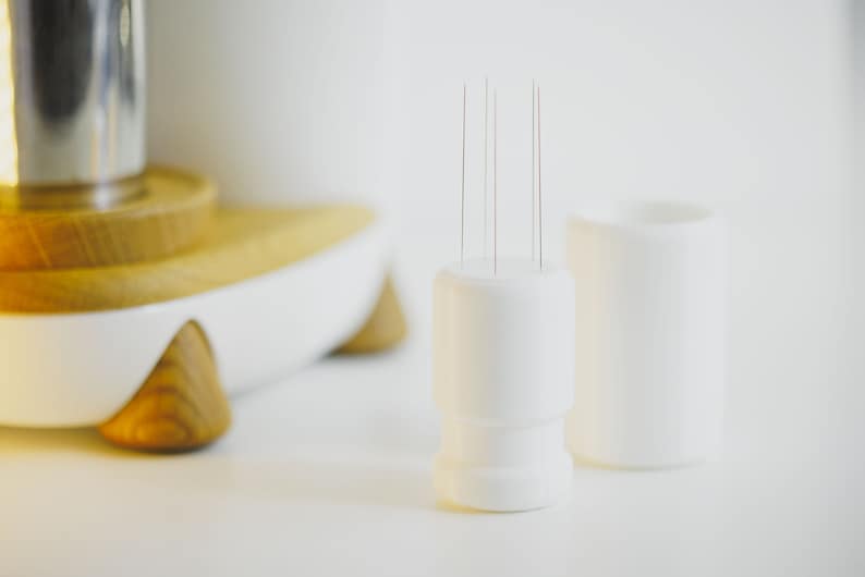 May include: A white ceramic holder with multiple thin needles sticking out of the top. The holder is sitting on a white surface next to another white ceramic holder.