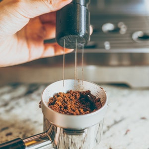 May include: A close-up of a silver portafilter filled with ground coffee beans. A black tamper with a needle-like tool is being held above the portafilter.