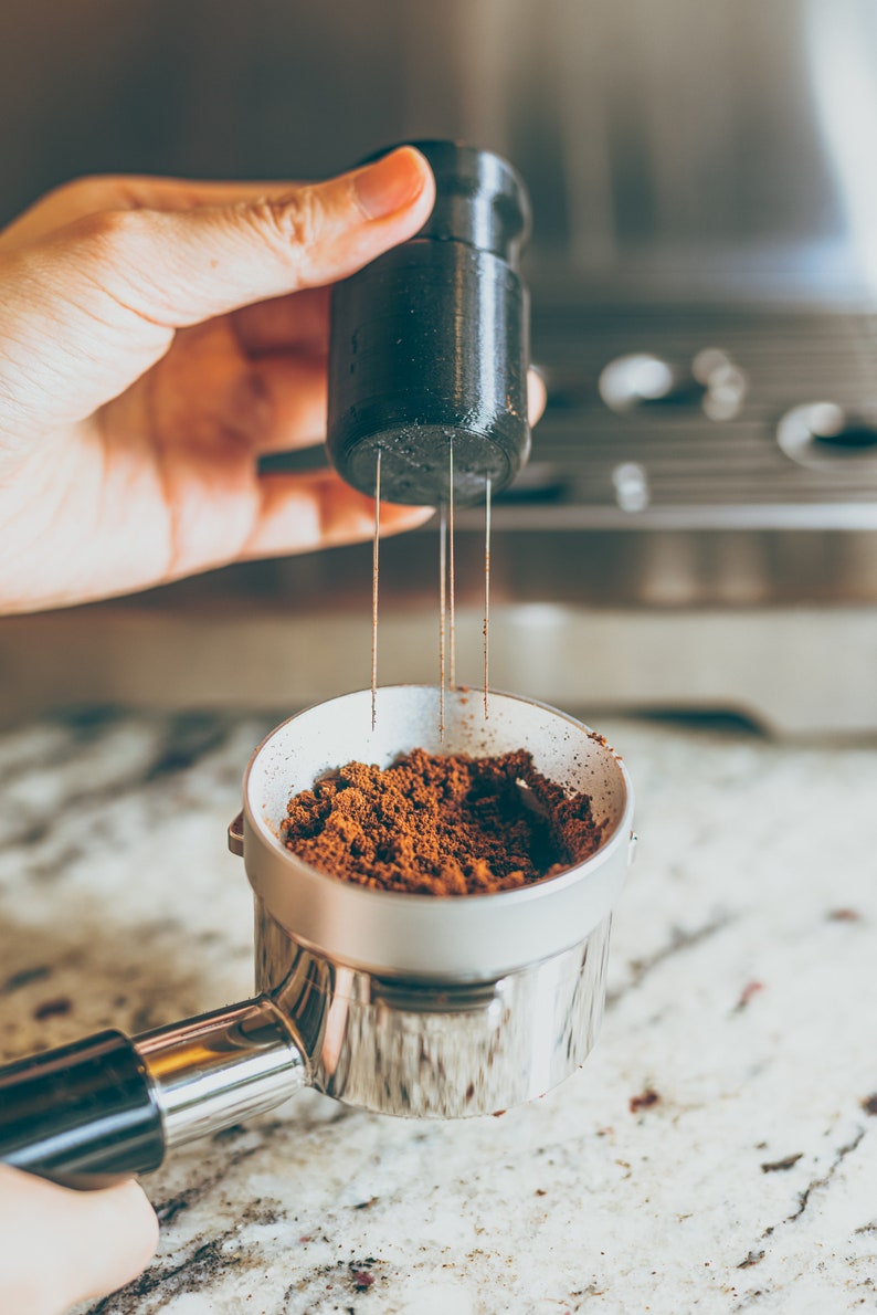 May include: A close-up of a silver portafilter filled with ground coffee beans. A black tamper with a needle-like tool is being held above the portafilter.