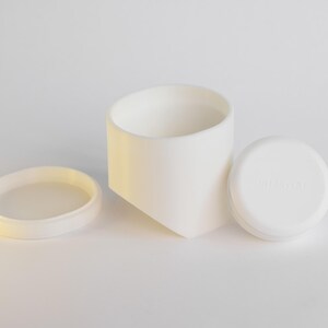 May include: A white ceramic container with a matching lid and a separate ring. The container has a geometric design, and the lid has text. The items are arranged on a white surface.