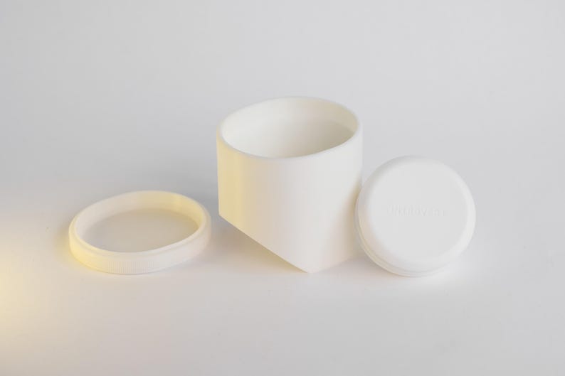 May include: A white ceramic container with a matching lid and a separate ring. The container has a geometric design, and the lid has text. The items are arranged on a white surface.