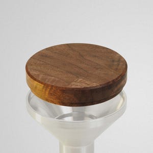 May include: A wooden lid with a circular design sits atop a silver funnel-shaped object. The lid is made of dark brown wood with visible grain. The words "FIVE OUNCE" and "INTROVERT" are printed in black.