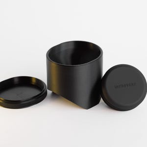 May include: A black container set with a cylindrical holder, a lid, and a base. The lid is round with the word "INTROVERT" printed on it. The base is a flat, circular disc with a ridged edge.