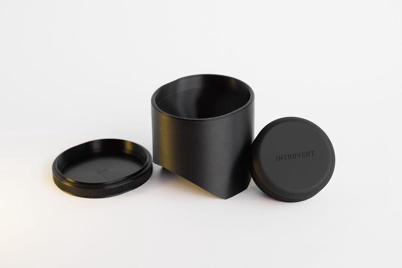 May include: A black container set with a cylindrical holder, a lid, and a base. The lid is round with the word "INTROVERT" printed on it. The base is a flat, circular disc with a ridged edge.
