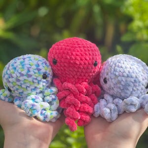 May include: Three handmade crochet octopus toys in shades of red, blue, and white. The plush toys have black button eyes and curled tentacles. The background is a blurred green.