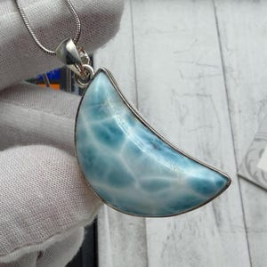 Può includere: Una collana con un ciondolo a forma di mezzaluna. Il ciondolo presenta una pietra preziosa azzurra con venature bianche, incastonata in una montatura in metallo argentato. La collana ha una delicata catena d'argento.