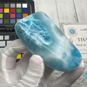 May include: A polished, oblong larimar gemstone, showcasing a blend of light blue and white hues with a unique, swirling pattern. The stone is held in a gloved hand, with a color calibration chart and a business card in the background.