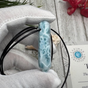 May include: A light blue larimar pendant with a black cord. The gemstone has white marbling and a polished finish. The pendant is held by a gloved hand. The background includes a red flower and a business card.