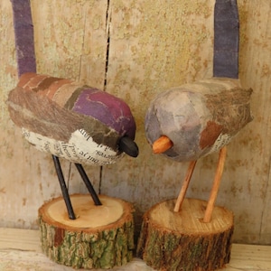 Può includere: Due figurine di uccelli fatti a mano. Un uccello ha la testa viola e l'altro grigia. Entrambi gli uccelli hanno corpi di carta e sono appollaiati su piccole basi di legno. Gli uccelli hanno zampe di legno e becchi neri o arancioni.