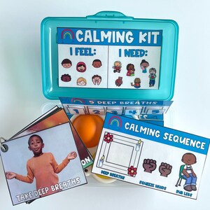 K&ouml;nnte beinhalten: Eine blaue Plastikbox mit einem Regenbogenmuster und dem Schriftzug "Calming Kit" auf dem Deckel. Die Box enth&auml;lt verschiedene Gegenst&auml;nde, darunter Karten mit Gesichtsbildern und den Worten "I Feel" und "I Need", eine Karte mit den Worten "Take Deep Breaths", eine Karte mit den Worten "Calming Sequence" und einen kleinen orangen Stressball.
