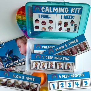 K&ouml;nnte beinhalten: Eine blaue Plastikbox mit einem Regenbogenmuster und dem Schriftzug "Calming Kit" auf dem Deckel. Die Box enth&auml;lt Karten mit Bildern und Text, darunter "Blow 5 Times", "5 Deep Breaths" und "I Feel: I Need:".