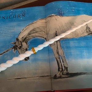 Op de afbeelding: Een witte eenhoornhoorn-toverstok met gouden accenten. De toverstok is gedraaid en heeft een afgeronde punt. De toverstok ligt op een boek met een tekening van een eenhoorn.