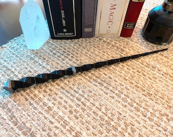 May include: A black and silver twisted wand with a pointed tip. The wand is lying on a beige textured surface.