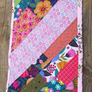 Op de afbeelding: Handgemaakte, gewatteerde tafelloper met een kleurrijk ontwerp. Het heeft diagonale strepen van bloemen- en geometrische patronen in roze, groen, blauw en oranje. De randen zijn afgewerkt met een roze bloemenrand, wat een decoratief stuk cre&euml;ert.