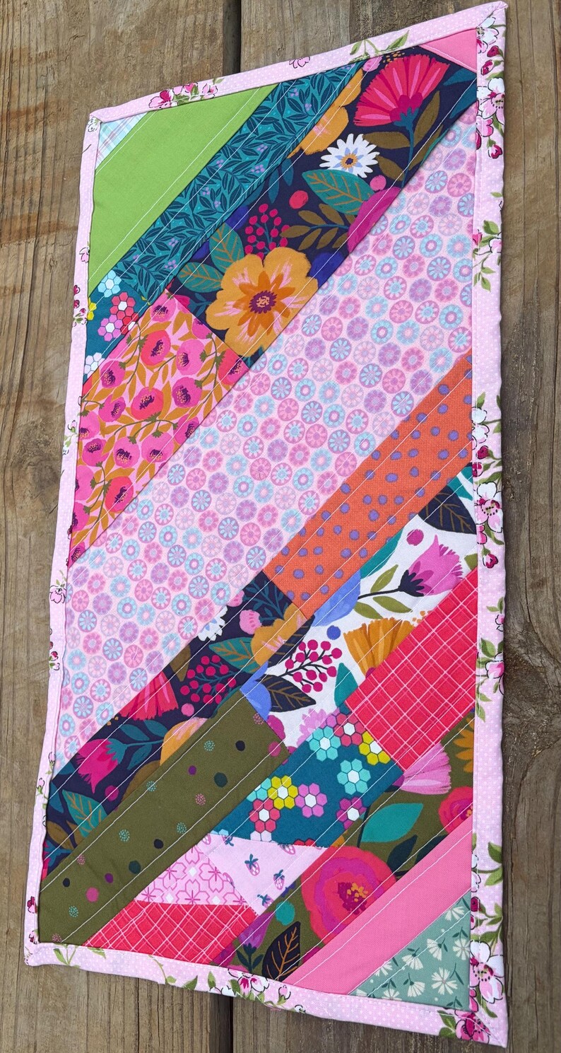 Op de afbeelding: Handgemaakte, gewatteerde tafelloper met een kleurrijk ontwerp. Het heeft diagonale strepen van bloemen- en geometrische patronen in roze, groen, blauw en oranje. De randen zijn afgewerkt met een roze bloemenrand, wat een decoratief stuk cre&euml;ert.