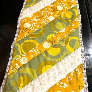 May include: A quilted table runner with a retro design. The runner features diagonal stripes of mustard yellow, olive green, and cream fabric. The yellow fabric has white floral patterns, while the green has large circles. The edges are finished with a patterned border.