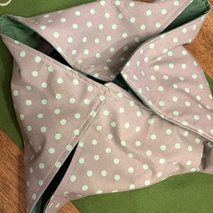May include: A gray fabric wrap with a polka dot pattern in light blue. The wrap is folded in a unique design, revealing a green interior. The wrap is on a green surface.