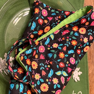 May include: A black fabric napkin with a vibrant floral and bird pattern, featuring pink, orange, and blue accents. The napkin has a bright green border and is tied with a matching strap. It rests on a clear glass plate.