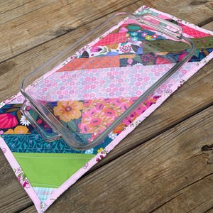 May include: A clear glass baking dish rests on a colorful patchwork quilted hot pad. The hot pad features a variety of floral and geometric patterns in pink, green, orange, and blue. The edges are finished with a pink binding.